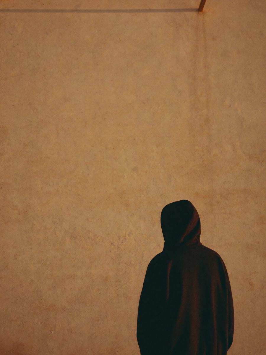 빈 벽에 대각선으로 떨어진 늦은 오후 햇빛, 그 아래 딥블랙 후디를 입고 등을 보인 인물